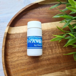 Könnte beinhalten: Eine kleine weiße Flasche mit einem blauen Etikett, auf dem "WAVE by Resin" steht. Die Flasche steht auf einem Holztablett.