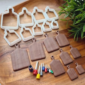 May include: A selection of white plastic cookie cutters in the shape of cutting boards, alongside brown wooden cutting boards of different sizes. There are also small metal tubes with coloured tips, all on a wooden tray.