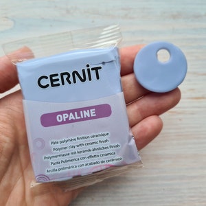 Może przedstawiać: Opakowanie jasnoniebieskiej gliny polimerowej z ceramicznym wykończeniem. Opakowanie jest oznaczone "CERNIT OPALINE" i zawiera instrukcje w wielu językach. Pokazano również mały, jasnoniebieski dysk z gliny.