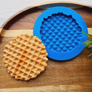 以下が含まれることがあります： ワッフル型のクッキーやお菓子を作るための青いシリコンモールド。モールドは、完成したワッフルクッキーの隣に置かれています。