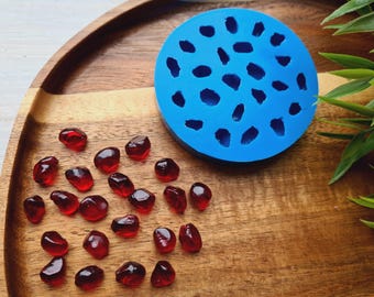 Puede incluir: Un molde de silicona azul con formas irregulares y varios objetos rojos translúcidos con forma de gema sobre una bandeja de madera. Se ven hojas verdes en el fondo. Los objetos se asemejan a semillas de granada.
