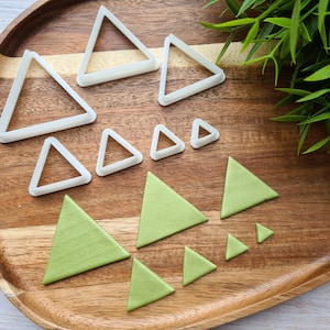 Pode incluir: Um conjunto de 10 cortadores de biscoitos de plástico branco em forma de triângulo e 10 formas de fondant de triângulo verde em uma bandeja de madeira.