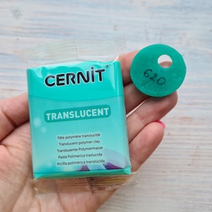 May include: A package of Cernit translucent polymer clay in a teal color, held in a hand. A teal-colored disc with a hole and the number "620" written on it is also visible. The package has text in multiple languages.