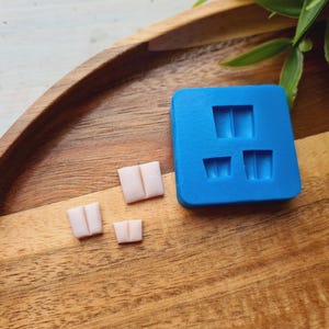 May include: A blue silicone mold with three rectangular cavities and three small, light pink, rectangular objects. The mold is square and sits on a wooden tray. The objects appear to be small soaps or candies.