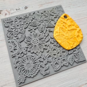 Puede incluir: Un sello de silicona gris con un diseño floral. Un molde de silicona amarillo con un diseño floral similar se coloca encima del sello.