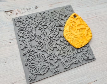 Pode incluir: Um carimbo de silicone cinza com um design floral. Um molde de silicone amarelo com um design floral semelhante é colocado em cima do carimbo.