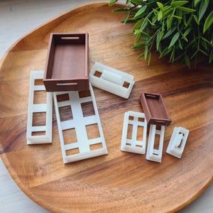 May include: A collection of miniature wooden trays and white plastic frames with rectangular cutouts. The trays are brown, and the frames are white. The items are arranged on a large, oval wooden tray, with green foliage in the background.