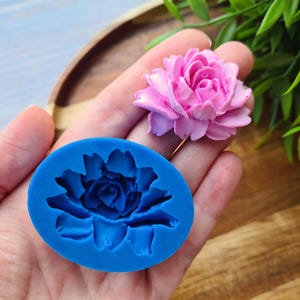 May include: A blue silicone mould shaped like a flower with a detailed design. The mould is being held in a hand, and a pink flower is visible in the background.