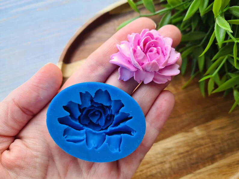 May include: A blue silicone mould shaped like a flower with a detailed design. The mould is being held in a hand, and a pink flower is visible in the background.