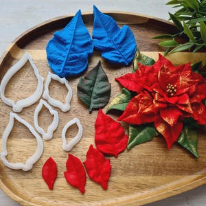 May include: A wooden tray holds a red poinsettia flower with gold accents, blue and green leaves, and white leaf-shaped cookie cutters. The poinsettia has a detailed texture, and the leaves are in various shades.