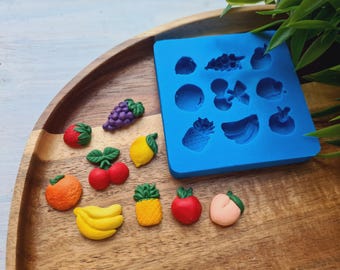 Puede incluir: Una colección de piezas de resina con forma de fruta y un molde de silicona azul. Las frutas incluyen uvas, fresas, limones, cerezas, naranjas, plátanos, piñas, manzanas y melocotones. El molde tiene formas de fruta para manualidades.