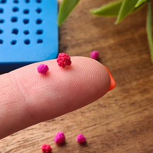May include: A close-up shot of tiny, handcrafted raspberries in shades of red and pink. The miniature berries sit on a fingertip, with a blue mould visible. These detailed sculptures suggest a craft project or miniature food creation.