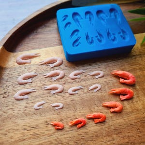 Op de afbeelding: Een blauwe siliconen mal met garnalenvormige holtes ligt op een houten dienblad, samen met verschillende kleine, roze garnalenvormige snoepjes en een paar oranje. De garnalensnoepjes variëren in grootte.