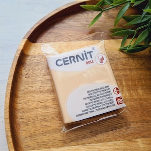 May include: A package of CERNIT DOLL polymer clay in a skin-tone color. The package is clear, showing the clay inside, and has white and red text. The package is on a wooden tray with green foliage.
