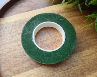 May include: A roll of green floral tape with a white inner core sits on a wooden surface. The tape is a solid, dark green color and appears to be new. The wooden surface has a natural wood grain pattern.