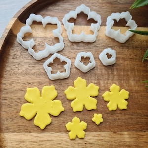 May include: A set of 10 white plastic flower cookie cutters and 6 yellow fondant flowers on a wooden surface. The cookie cutters are in various sizes and shapes.