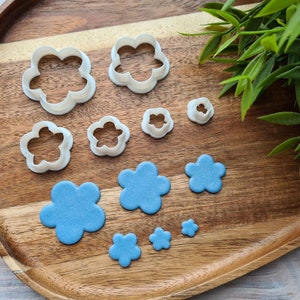 Puede incluir: Un juego de seis cortadores de galletas con forma de flor de varios tamaños, hechos de plástico blanco, sobre una superficie de madera. También hay seis flores de fondant azules, cortadas con los cortadores de galletas.