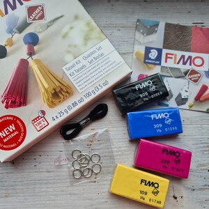 May include: A Fimo Tassel Kit with four 25 gram blocks of polymer clay in black, blue, pink, and yellow. The kit also includes black string, silver jump rings, and instructions for making tassels. The box reads "Tassel Kit - Quasten Set - Kit Tassels - Set Borlas".
