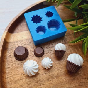 Puede incluir: Un molde de silicona azul con cuatro cavidades en forma de cupcake, junto con seis mini cupcakes, tres con glaseado de chocolate y tres con glaseado blanco.