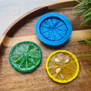 Puede incluir: Tres moldes de silicona para hacer manualidades de resina. Los moldes tienen forma de rodajas de fruta, incluyendo una lima, un limón y una naranja. Los moldes están sobre una superficie de madera.