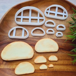 Op de afbeelding: Een verzameling witte plastic koekjessnijders in verschillende vormen, waaronder boogvormige ramen en afgeronde vormen, samen met verschillende gebakken koekjes. De uitstekers zijn gerangschikt op een houten dienblad, met een takje groene bladeren op de achtergrond.