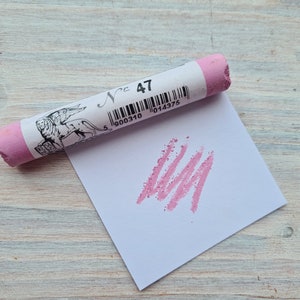 Puede incluir: Un palo de pastel rosa con una etiqueta blanca que dice "N° 47" y un código de barras. El palo está sobre un papel blanco con una línea de pastel rosa.