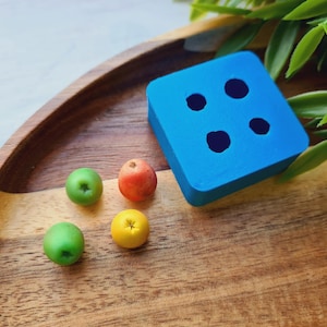 Peut inclure: Un plateau en bois présente un carré bleu avec quatre cercles foncés et plusieurs petits objets en forme de fruits colorés. Deux fruits verts, un orange et un jaune sont dispersés sur le plateau, avec un feuillage vert en arrière-plan.