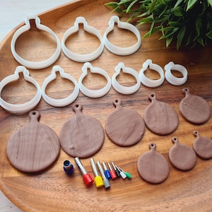 Peut inclure: Un ensemble de découpoirs à biscuits blancs en forme de boules de Noël et d'ornements en argile marron de différentes tailles, disposés sur un plateau en bois. Plusieurs petits outils avec des manches colorés sont également présents. L'ensemble est sur un fond de verdure.