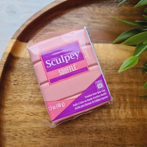 Puede incluir: Un paquete de arcilla polimérica Sculpey Soufflé de color rosa claro. El paquete tiene una etiqueta morada y rosa con el nombre de la marca y el tipo de producto. Pesa 48 g.