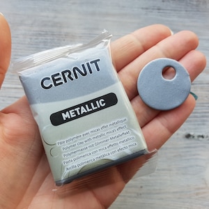 Puede incluir: Un paquete de arcilla polim&eacute;rica met&aacute;lica plateada de la marca Cernit. El paquete tiene texto en varios idiomas que describe el producto. Se muestra un peque&ntilde;o disco plateado con un agujero en el centro junto al paquete.