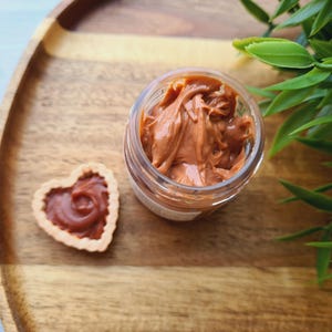 May include: A small glass jar filled with caramel-coloured cream sits on a wooden tray. Beside it is a heart-shaped pastry filled with the same cream. Green leaves are in the background.