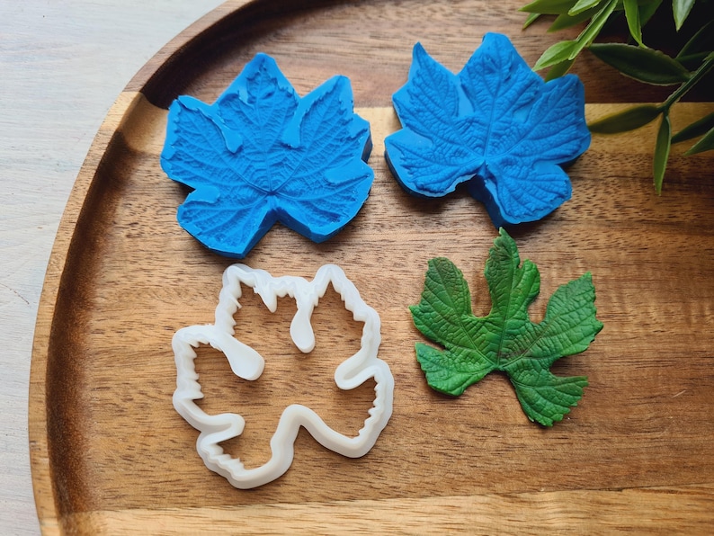 May include: Four leaf-shaped cookie cutters, two blue, one white, and one green. The cutters are on a wooden surface.