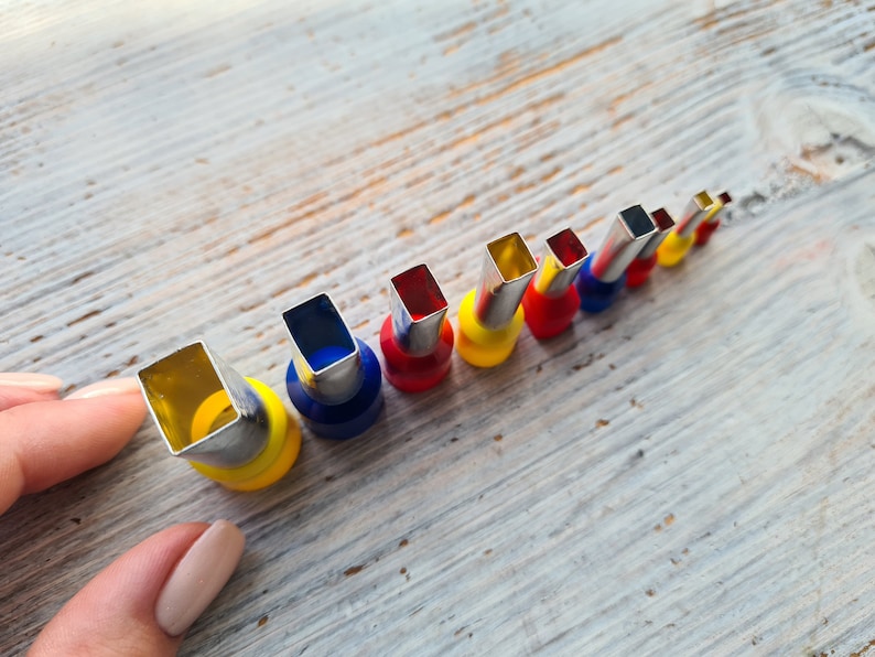 May include: A set of colorful crimping tools with rectangular heads, arranged in a diagonal row. The tools have silver metal heads and are accented with yellow, blue, and red bases. The tools are on a light-colored wooden surface.