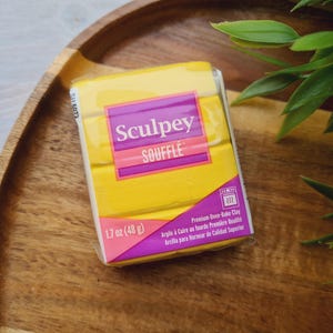 May include: A package of yellow Sculpey Soufflé polymer clay, 1.7 oz (48 g), with a pink and purple label. The package is on a wooden tray with green leaves in the background. The text on the package says "Premium Oven-Bake Clay".