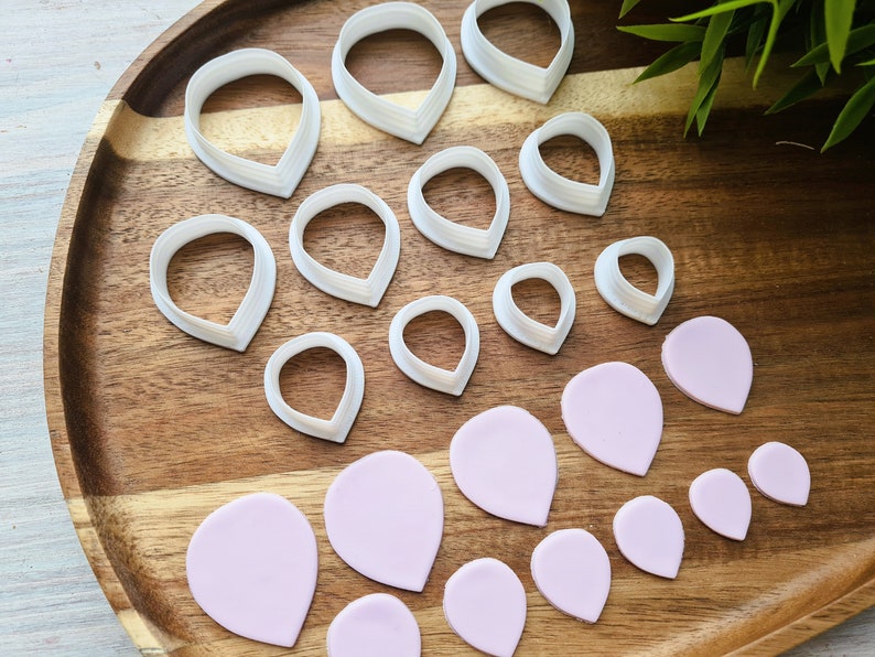 Może przedstawiać: Zestaw 12 białych plastikowych foremek do wycinania płatk&oacute;w kwiat&oacute;w i 12 r&oacute;żowych płatk&oacute;w kwiat&oacute;w z masy cukrowej na drewnianej tacy.