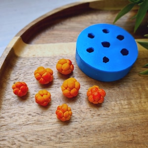 May include: A blue silicone mold with seven round cavities for making small, orange-colored, cloudberry-shaped candies. The mold is on a wooden surface with several cloudberries scattered around it.
