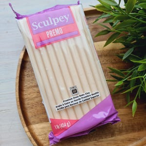 Könnte beinhalten: Eine Packung Sculpey Premo Polymer Clay, 454 g, in einer transparenten Kunststoffverpackung mit einem lila und rosa Etikett. Die Packung enthält mehrere beige Tonstäbchen. Der Text "Premium Oven-Bake Clay" ist sichtbar.