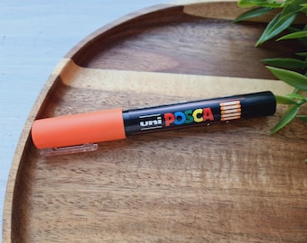 May include: Orange and black Uni Posca paint marker with a white cap. The marker is on a wooden surface.