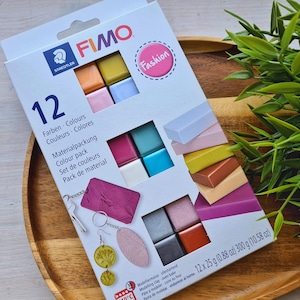 Op de afbeelding: Een doos FIMO boetseerklei in 12 kleuren, met een "Fashion" label. De doos bevat 12 kleurstaven van elk 25g, in totaal 300g. De doos staat op een houten dienblad met groen gebladerte.
