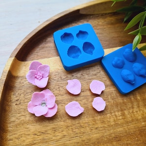 Puede incluir: Dos moldes de silicona azul para hacer flores de arcilla polimérica. Un molde tiene cuatro formas de pétalos de flores, y el otro tiene tres formas de pétalos de flores más grandes. También hay seis pétalos de flores de arcilla polimérica rosa y dos flores de arcilla polimérica rosa.