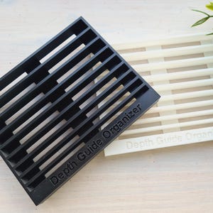 Peut inclure: Deux organisateurs rectangulaires &agrave; fentes, l'un noir et l'autre blanc. Le texte "Depth Guide Organizer" est imprim&eacute; sur les deux. Ces organisateurs sont con&ccedil;us pour ranger et organiser des objets.