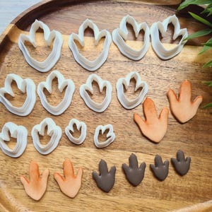 Puede incluir: Una colección de cortadores de galletas y recortes de fondant con forma de huellas de dinosaurio en varios tamaños. Los cortadores son blancos y el fondant es de tonos naranja y marrón, dispuestos sobre una bandeja de madera.