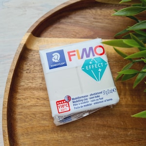 May include: A white package of FIMO Effect modeling clay with a blue and white diamond design. The package has the text "STAEDTLER" and "FIMO EFFECT" on it. The package also includes instructions for using the clay, including the baking temperature of 110°C (230°F).