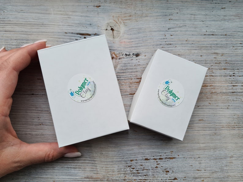 May include: Two white rectangular boxes with rounded corners, one larger than the other, are displayed. Each box has a circular sticker with the text "Polymer Clay" and a website address. The boxes are on a light wood surface.