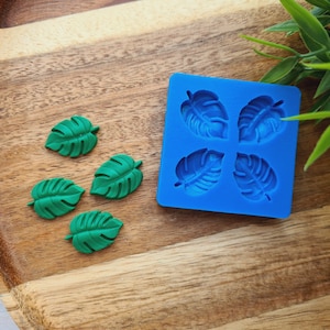 Puede incluir: Un molde de silicona azul con cuatro formas de hojas, y cuatro piezas de resina en forma de hojas verdes sobre una superficie de madera.
