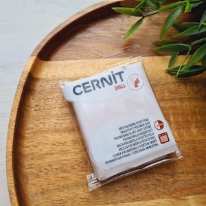 May include: A package of Cernit Doll polymer clay in a clear plastic wrapper, resting on a wooden tray. The package has text in multiple languages, including "Skin Effect Polymer Clay". Green leaves are in the background.