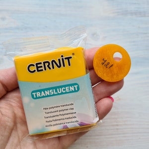 May include: A yellow package of translucent polymer clay by Cernit. The package has the brand name and product name printed on it in black text. The package is sitting on a person's hand. A small yellow circle with the number 421 written on it is also visible in the image.