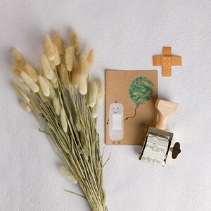 Puede incluir: Una tarjeta marr&oacute;n con un personaje de dibujos animados sosteniendo una venda y un &aacute;rbol verde sobre un fondo blanco. Un sello de goma con la palabra "freuden" est&aacute; al lado de la tarjeta. Un mont&oacute;n de hierba beige seca est&aacute; en el fondo.