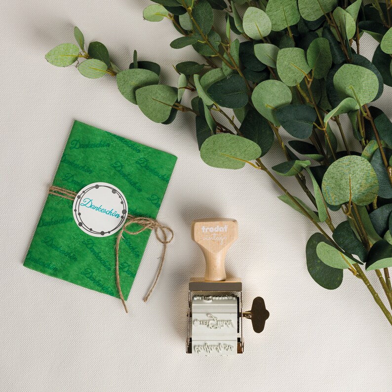 Puede incluir: Un sobre de papel verde con el texto "Danke sch&ouml;n" en una pegatina circular blanca. Un sello de madera con el texto "trodat vintage" est&aacute; al lado del sobre. El sello tiene una base de metal y un mango con una perilla de lat&oacute;n.