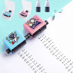 Könnte beinhalten: Ein blau-rosa rollender Mathematikstempel mit Cartoon-Astronauten- und Charakterdesigns. Der Stempel wird verwendet, um Matheaufgaben auf Papier zu erstellen. Zwei Tintenflaschen und zwei transparente Kunststoffgehäuse sind ebenfalls sichtbar.